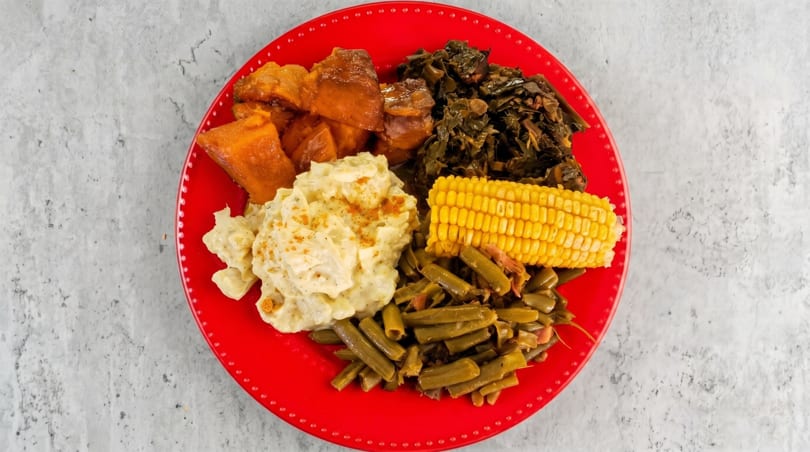 A red plate filled with candied yams, collard greens, potato salad, corn on the cob, and green beans.