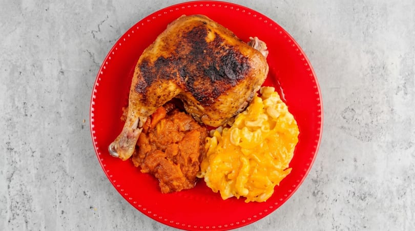A red plate holds a roasted chicken leg quarter, mashed sweet potatoes, and macaroni and cheese.